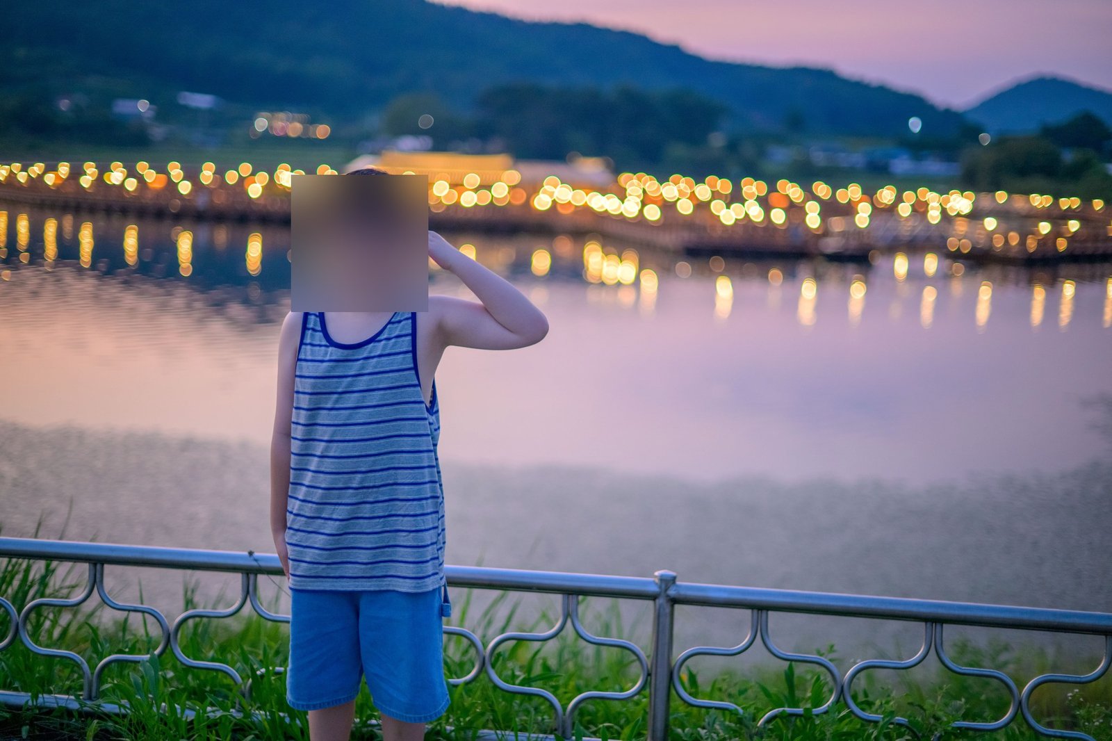 해 질 녘 호수와 다리 조명의 보케를 배경으로 경례하는 아이(얼굴 블러 처리)