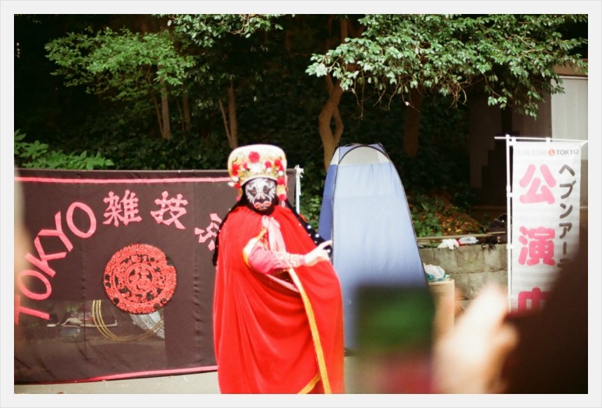 붉은 망토와 금색 모자, 얼굴 가면을 쓴 공연자가 TOKYO 雑技라고 적힌 검은 현수막 앞에 서 있다