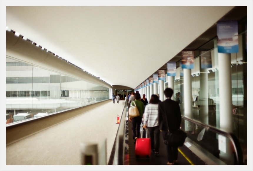 공항 안의 긴 통로, 무빙워크 위로 사람들이 멀어져 가고 붉은 캐리어가 보인다