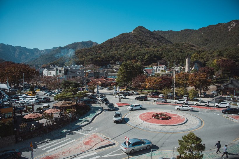로터리와 횡단보도 주변 도로의 승용차와 보행자, 뒤쪽에 단풍이 든 산비탈