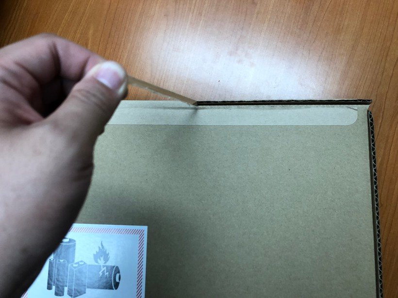 한 손으로 갈색 박스 윗변의 뜯는 띠를 잡아당기는 모습