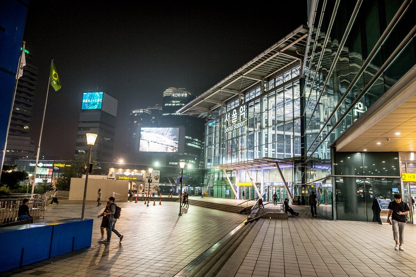 밤의 서울역 앞 광장. 유리 외벽에 서울역 Seoul Station 글자가 켜져 있고 광장에 가로등과 보행자가 보인다