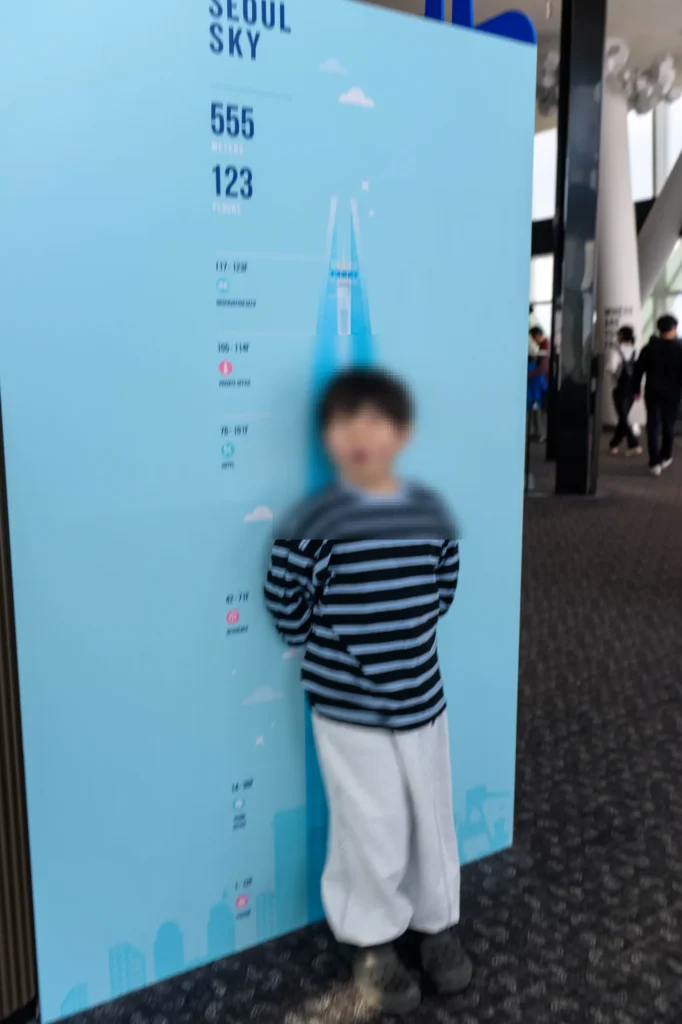 SEOUL SKY 555 METERS 123 FLOORS가 적힌 하늘색 키재기판 앞에 선 아들