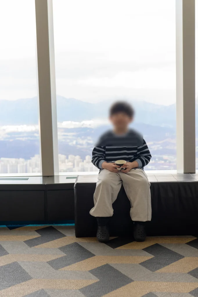전망대 창가 검은 벤치에 앉아 컵을 든 아들, 뒤로 흐릿한 산과 도시