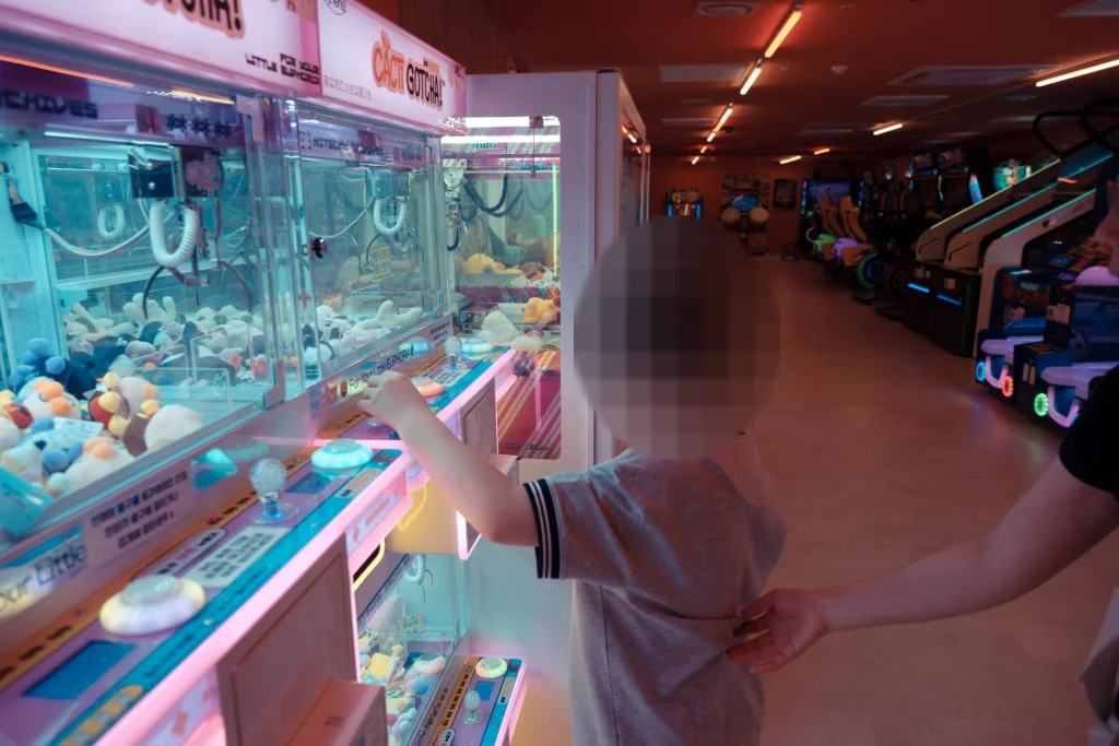 숙소 안 오락실 인형뽑기 기계 앞에서 버튼을 누르는 아이와 아이의 옷자락을 잡은 손