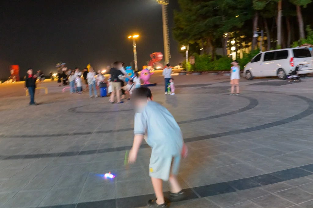 해변 광장에서 바닥의 빛나는 장난감을 쫓는 아이, 흔들린 사람들