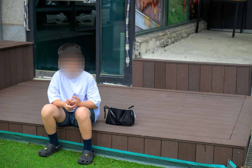 가게 앞 나무 데크에 앉아 두 손을 모은 아이와 옆에 놓인 검은 가방