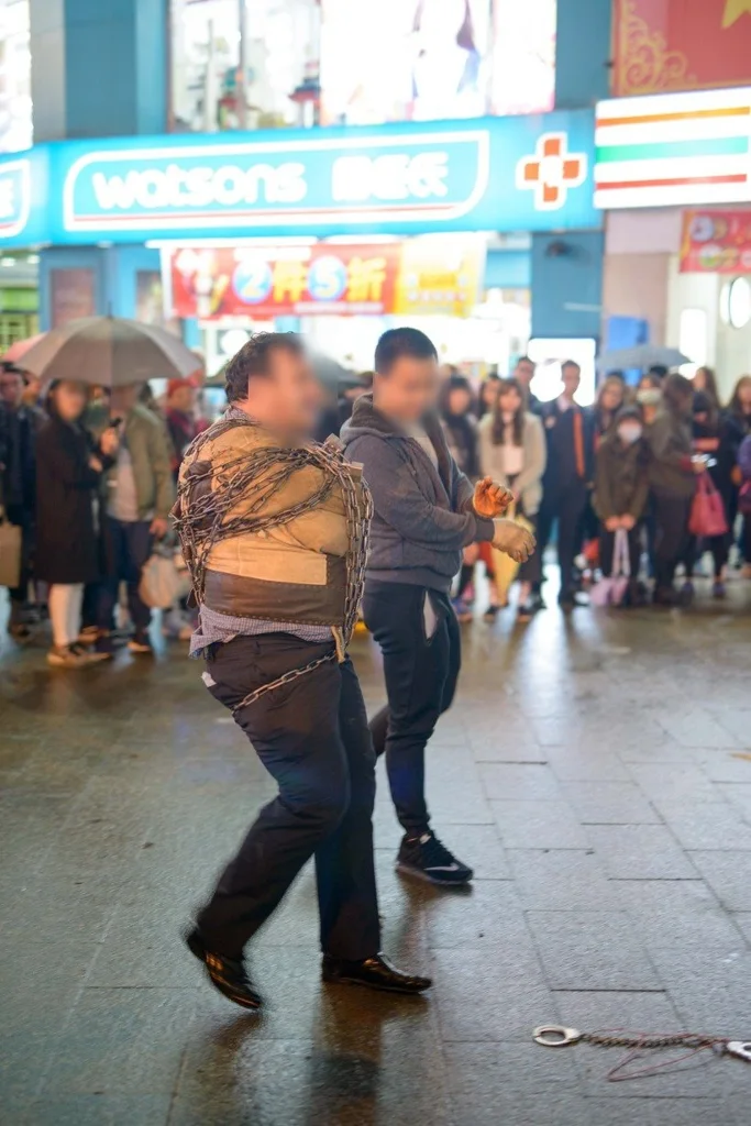 몸에 사슬처럼 보이는 줄이 감긴 사람과 다른 한 사람이 서 있고 뒤에 watsons 간판이 보이는 밤거리 (세로 사진, 얼굴 가림)