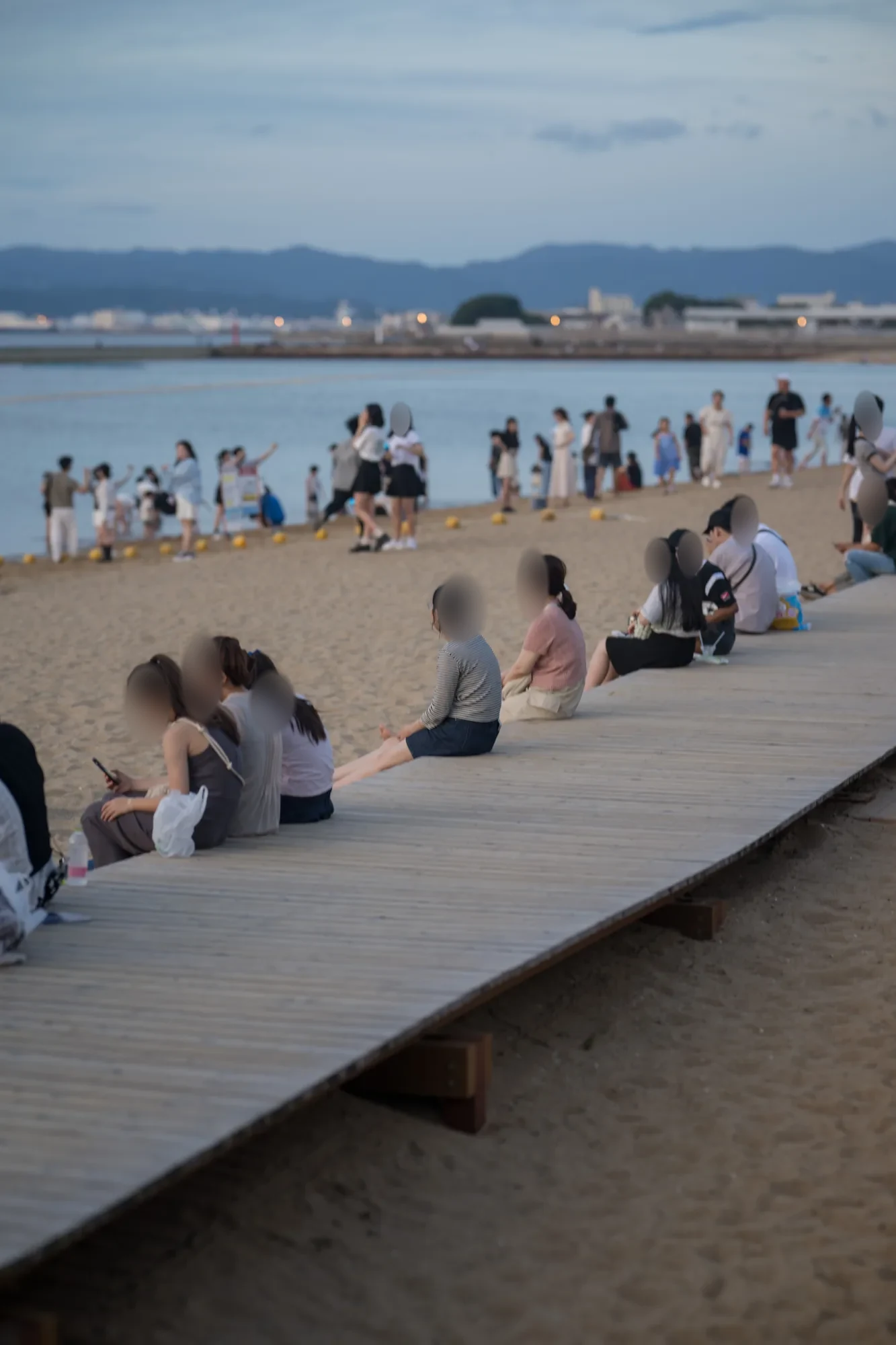 모래사장 옆 나무 데크에 줄지어 앉은 사람들과 물가의 노란 부표, 얼굴은 모자이크로 가렸다