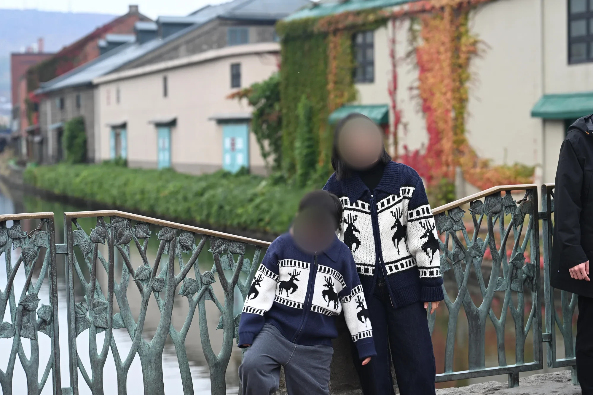 잎 모양 장식이 달린 난간 앞에서 같은 무늬의 카디건을 입은 어른과 아이, 얼굴은 흐림, 뒤로 운하와 담쟁이 덮인 건물