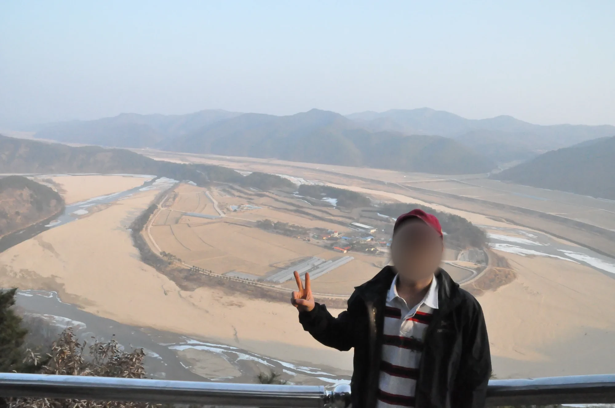 전망대 난간 앞에서 손가락으로 브이를 한 빨간 모자 차림의 남자, 얼굴은 흐림