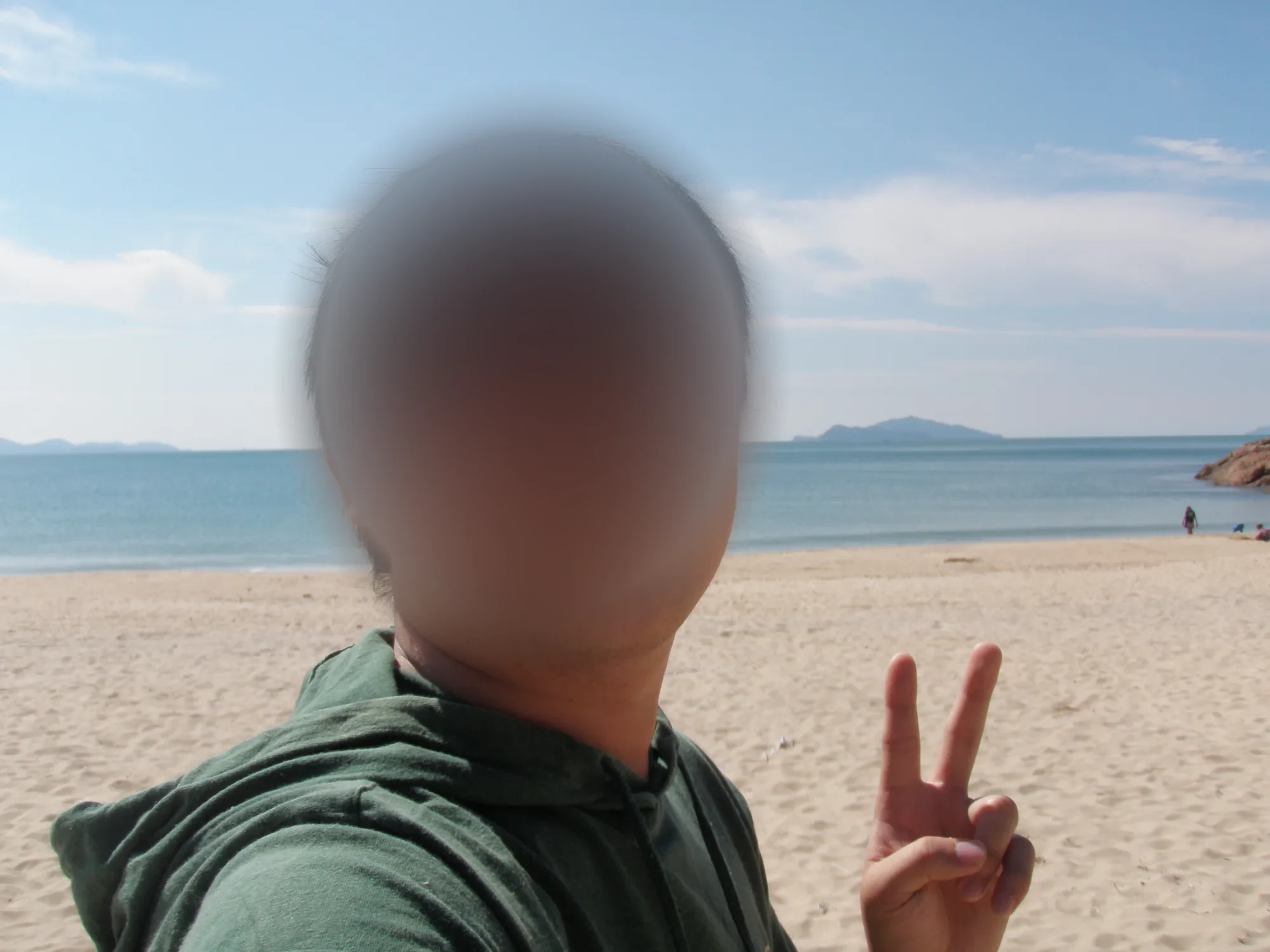 모래사장과 바다를 배경으로 초록색 후드 점퍼를 입은 안경 쓴 남자가 브이 손짓을 한 모습, 얼굴은 흐림 처리