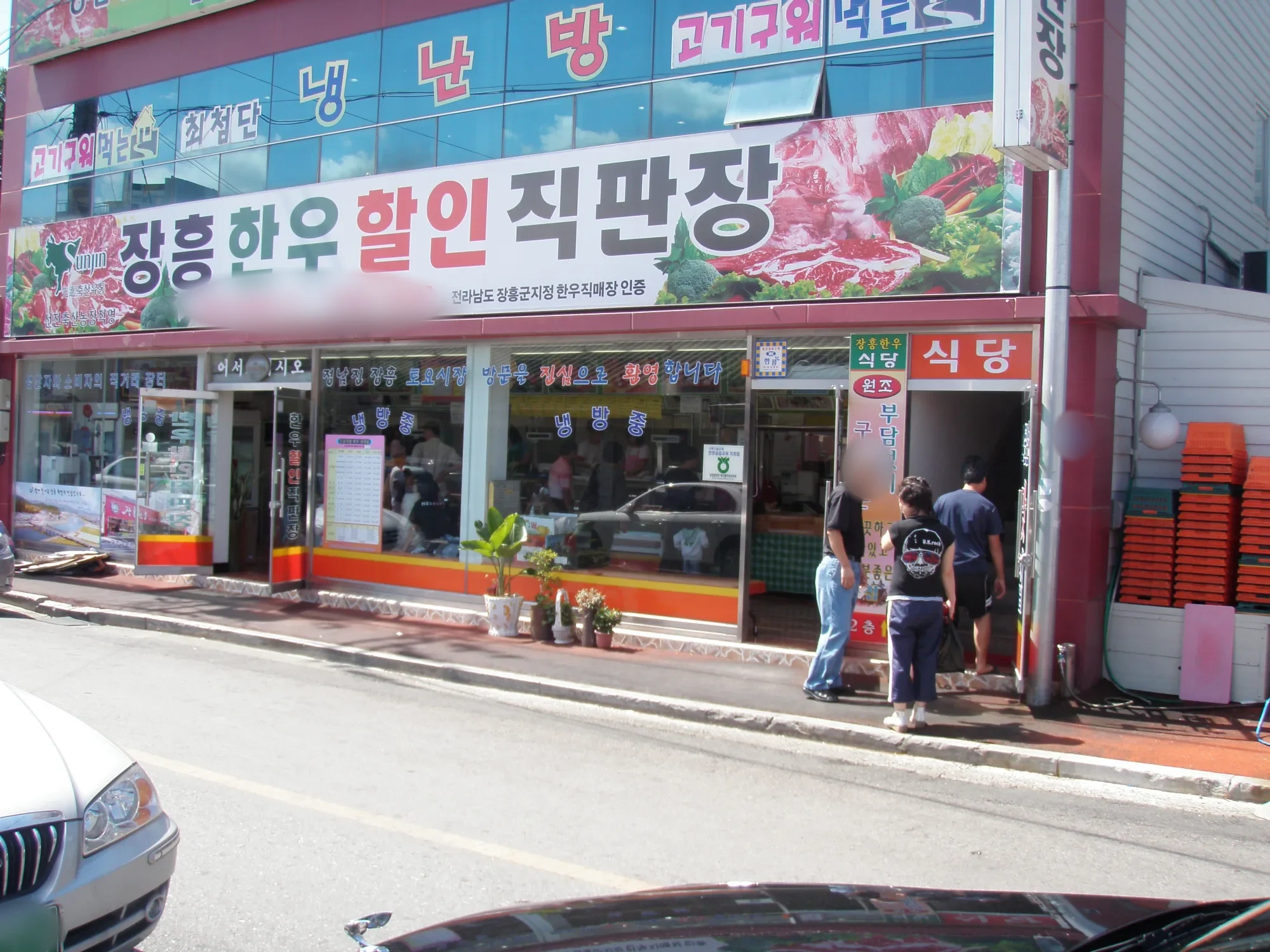붉은 고기 그림이 있는 큰 간판의 상점 앞 도로와 출입구 앞에 선 세 사람, 얼굴과 전화번호와 번호판은 흐림 처리