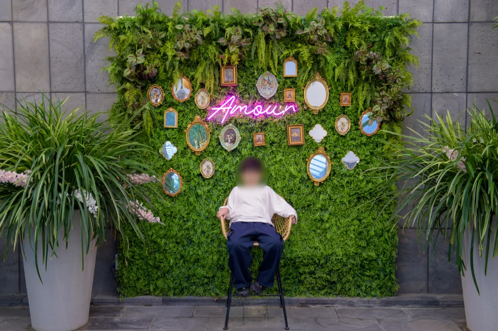 거울과 액자가 걸린 초록 식물 벽 앞 의자에 앉은 아들