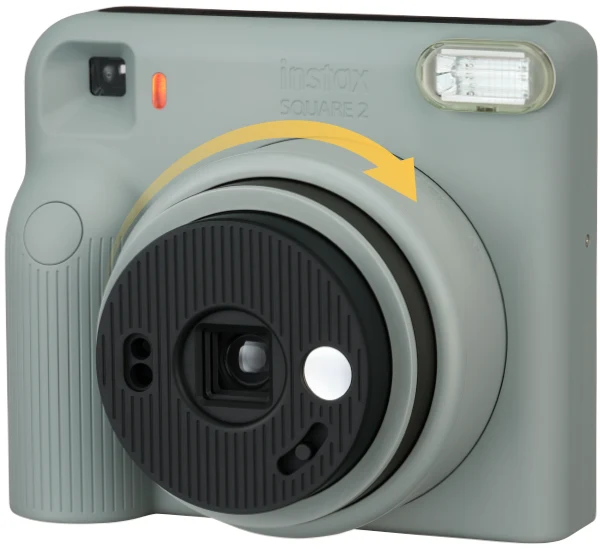 instax SQUARE 2 청회색 몸체를 비스듬히 본 모습, 렌즈 링을 시계 방향으로 돌리는 노란 화살표 표시