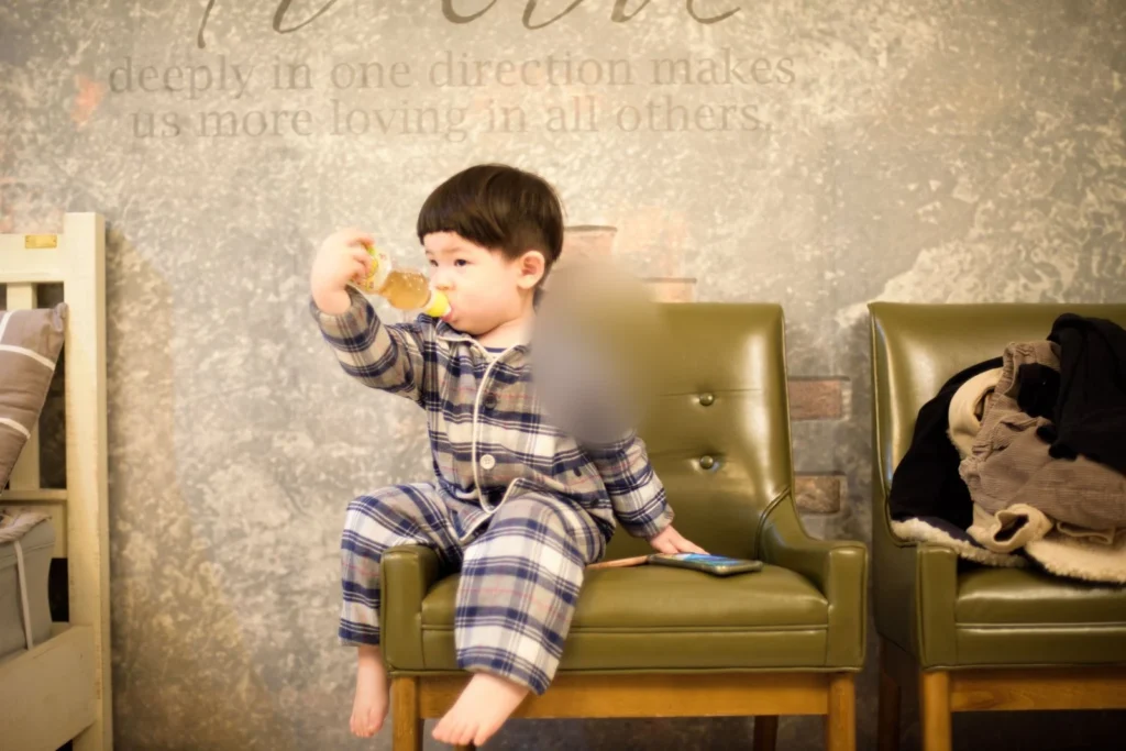 녹색 가죽 의자에 앉아 병을 위로 든 아이, 뒤쪽 벽에 영문이 적혀 있다(얼굴 가림)