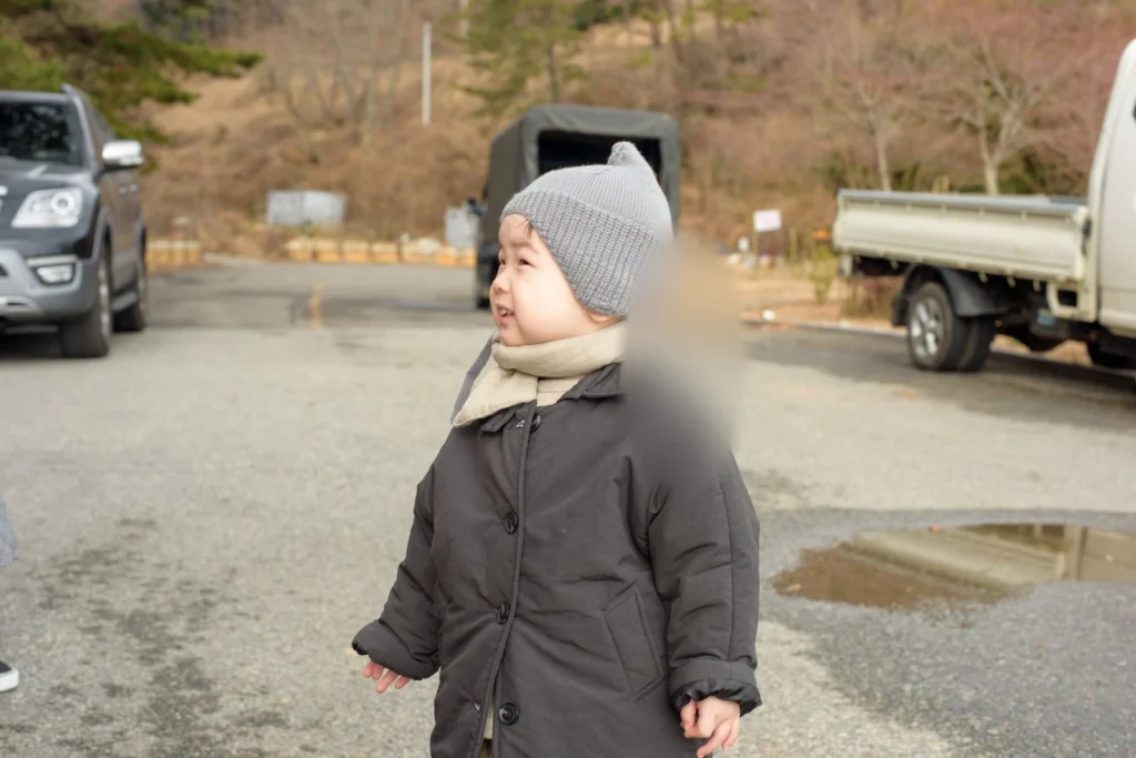 주차장에서 회색 니트 모자와 어두운 색 패딩을 입고 선 아이, 뒤쪽에 승용차와 트럭이 있다(얼굴 가림)
