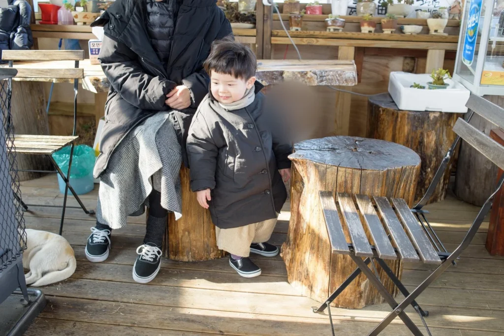 나무 데크에서 어른 옆에 선 아이, 왼쪽 아래에 흰 개가 누워 있다(얼굴 가림)