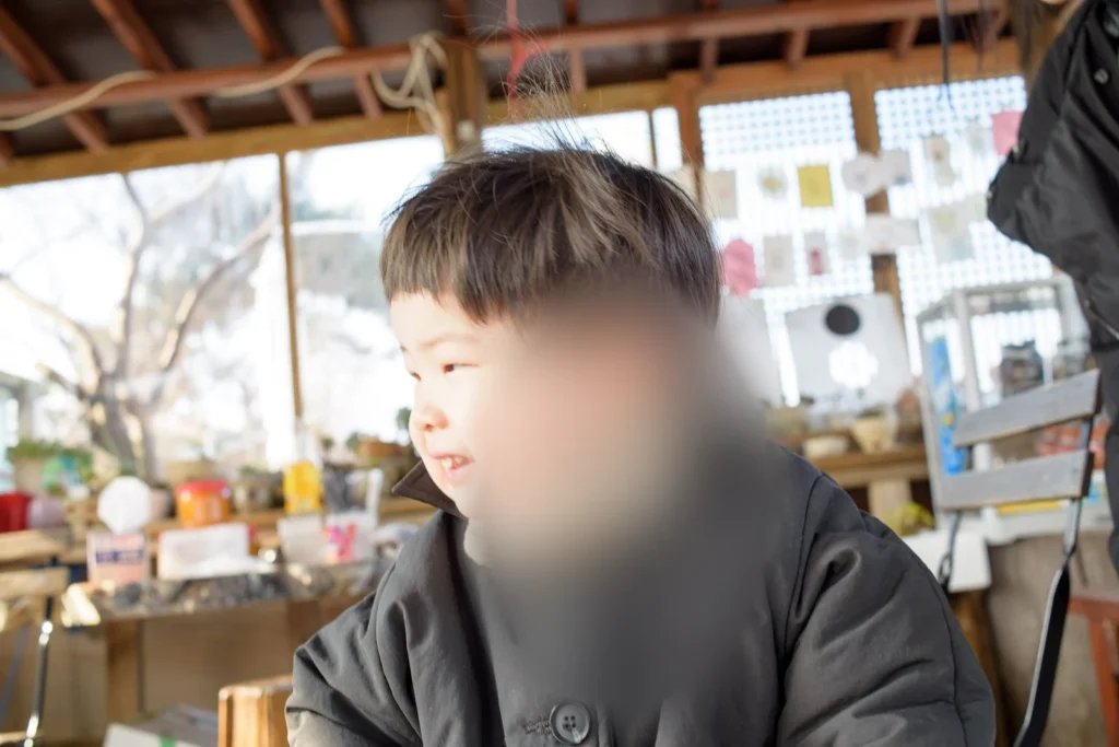 창가에서 고개를 왼쪽으로 돌린 아이, 뒤쪽 선반과 창에 작은 물건이 있다(얼굴 가림)