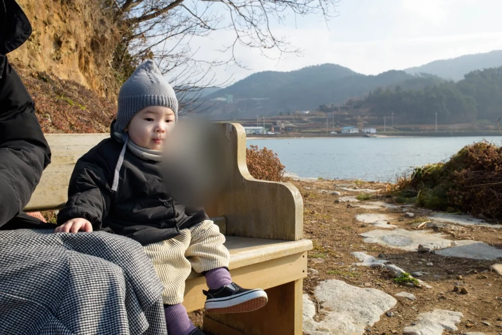 바닷가 나무 의자에 앉은 아이, 왼쪽 아래에 어른의 무릎 담요가 보인다(얼굴 가림)