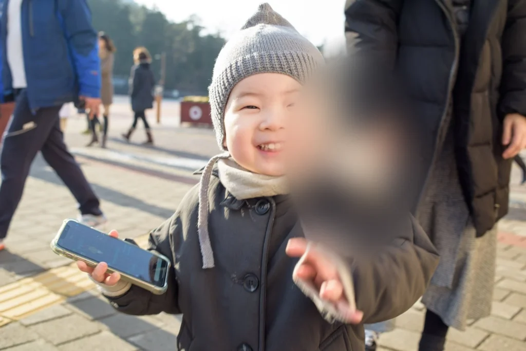 스마트폰을 든 아이, 뒤쪽에 어른과 걸어가는 사람들이 보인다(얼굴 가림)