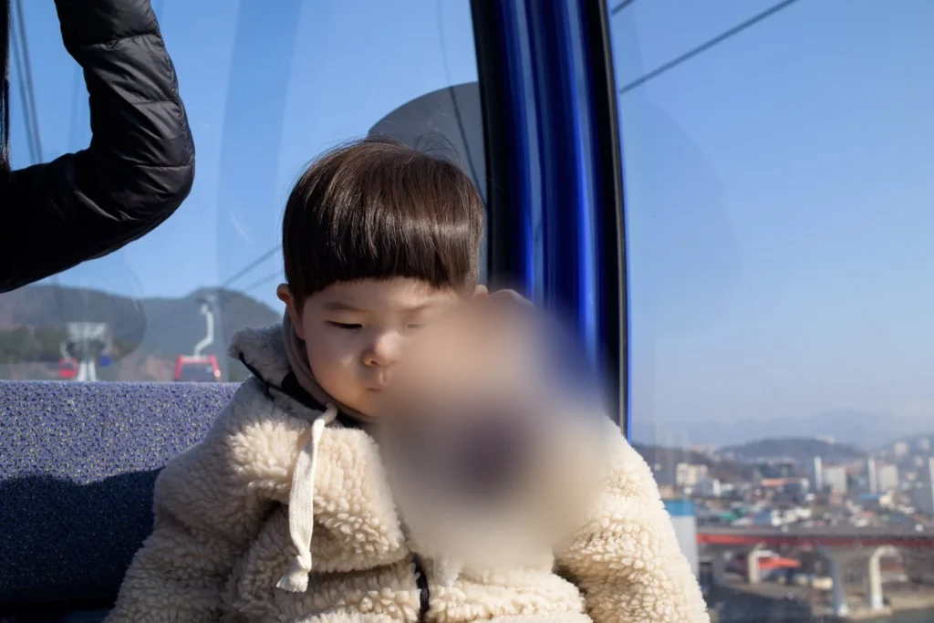 파란 창틀 옆 좌석에 앉은 아이, 창밖에 도시가 보인다(얼굴 가림)
