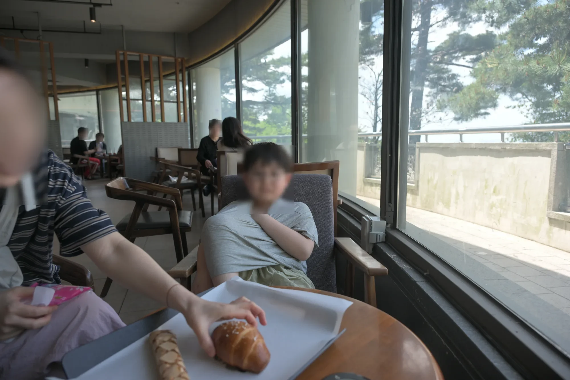 창가 의자에 앉은 아이와 탁자 위 쟁반에 놓인 빵, 왼쪽에서 손을 뻗는 사람(얼굴 모자이크)
