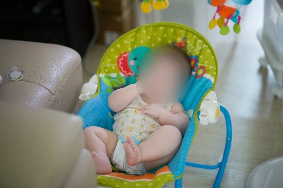 거실 바닥의 알록달록한 바운서 의자에 앉아 손을 입에 댄 아기, 얼굴을 가림