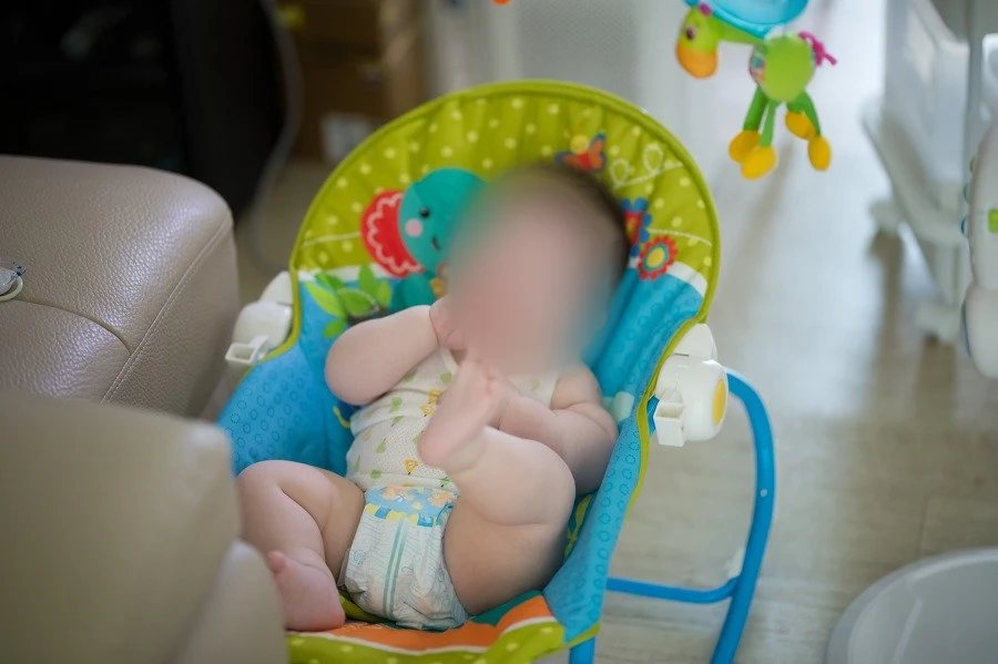 바운서 의자에 앉아 두 손을 입 가까이 대고 한쪽 발을 든 아기, 얼굴을 가림