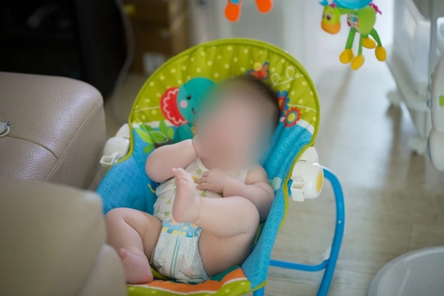 바운서 의자에 앉아 손을 입 가까이 대고 한쪽 발을 든 아기, 얼굴을 가림