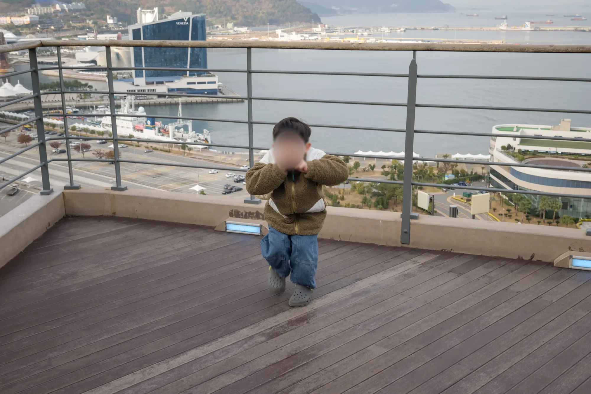 나무 데크 전망대에서 손을 얼굴 앞에 올린 아이, 뒤로 항구와 시가지가 보임, 얼굴을 가림