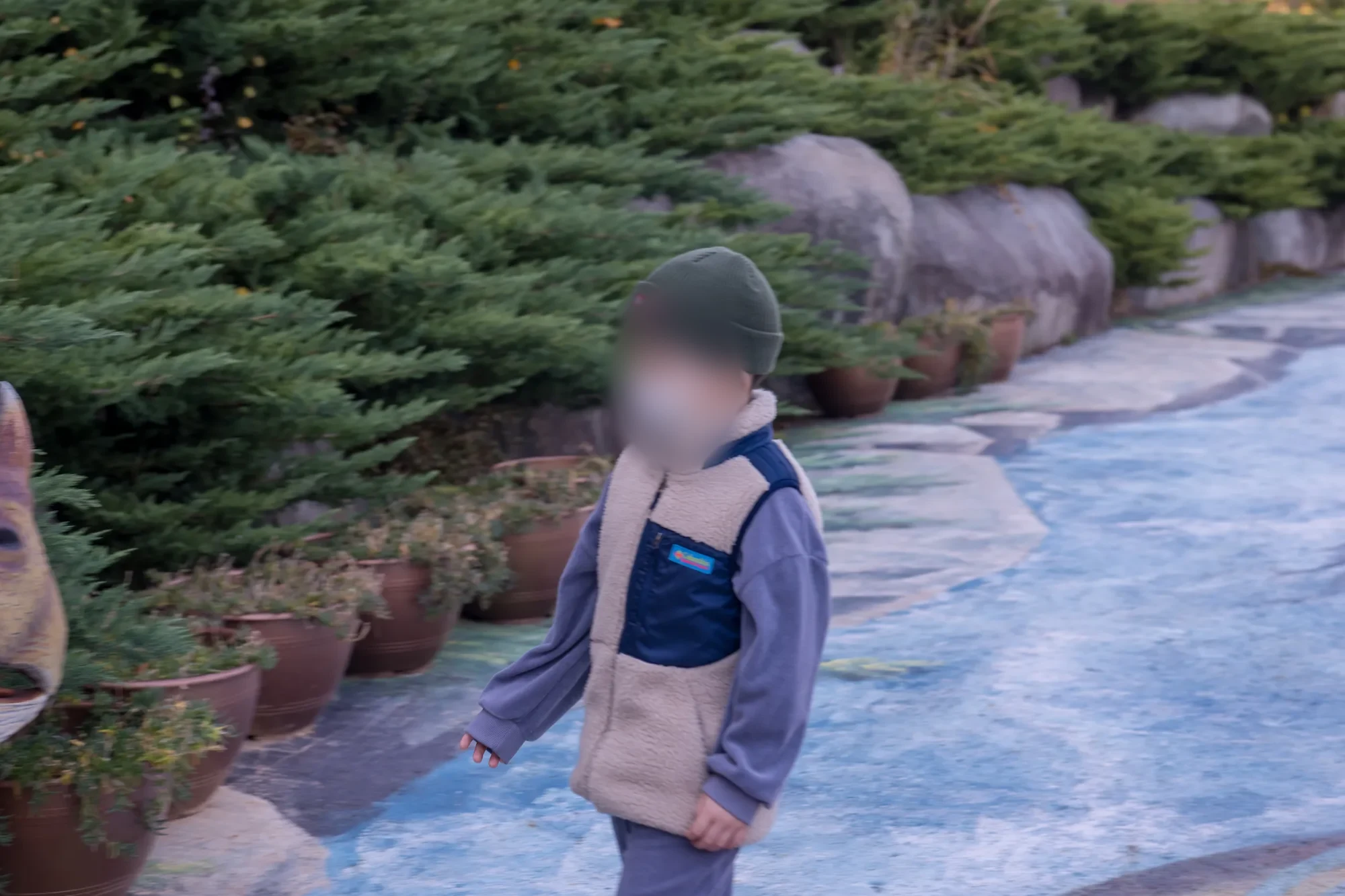 바위 정원 옆 파란 바닥 길에 마스크를 쓰고 서 있는 아이, 얼굴을 가림